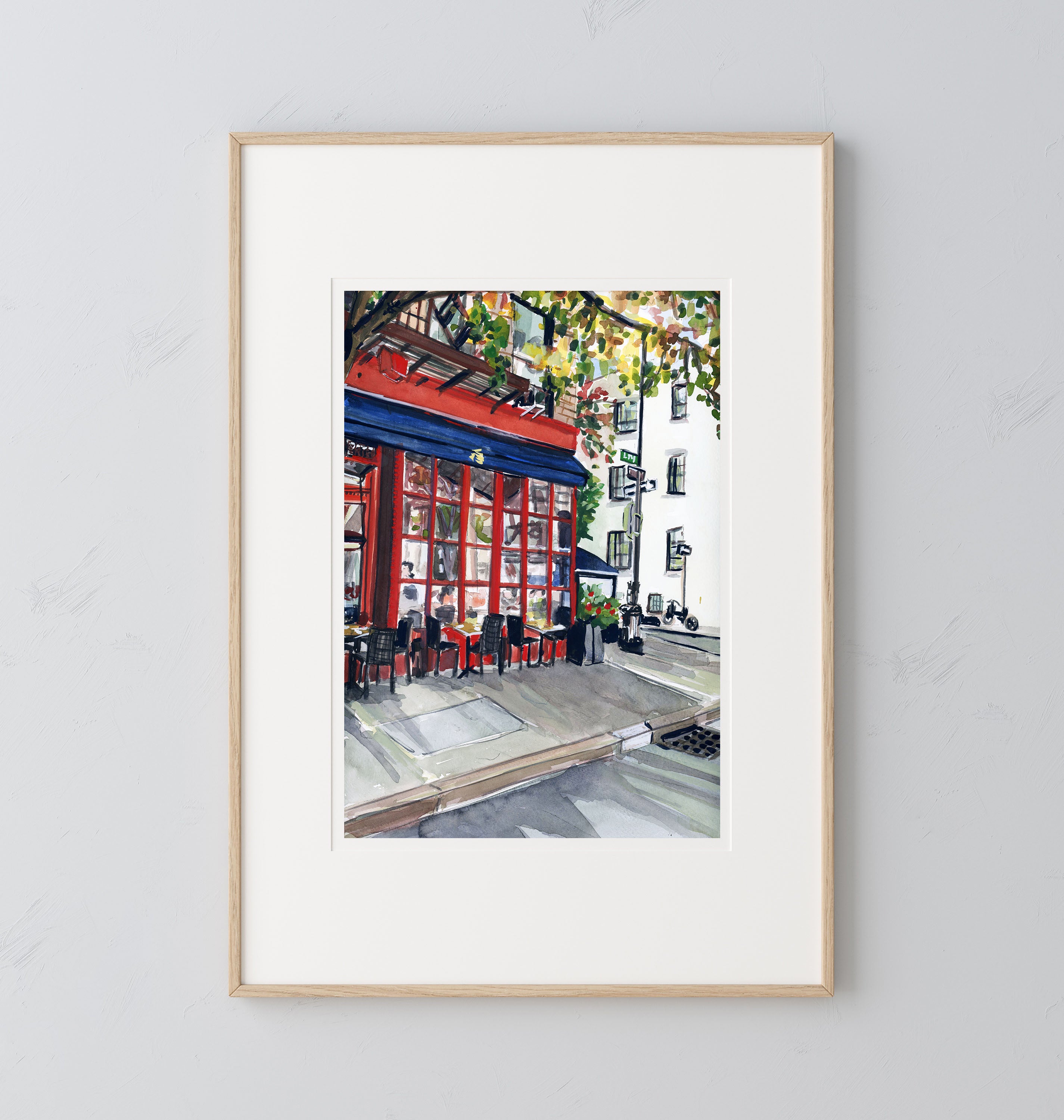 NYC Cafe print of painting by Medjool Studio. Print of original gouache painting showing a vibrant red cafe that stands out against the muted tones of the surrounding buildings. This print also shows glimpses of the cozy interior, with patrons seated at tables adorned with fresh flowers, their faces softly illuminated by the warm glow of the café's interior lights.
