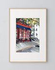 NYC Cafe original painting by Medjool Studio. Original gouache painting showing a vibrant red cafe that stands out against the muted tones of the surrounding buildings. This painting also shows glimpses of the cozy interior, with patrons seated at tables adorned with fresh flowers, their faces softly illuminated by the warm glow of the café's interior lights.